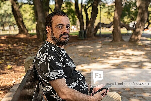 Ein Mann sitzt auf einer Bank im Park mit seinem Handy in der Hand und schaut in die Kamera