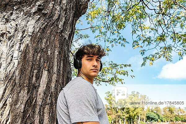 A young man listening to music  leaning on a tree with his arms folded  and looking at camera