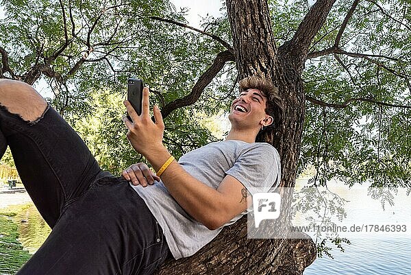 Junger Mann lehnt an einem Baumstamm  lacht und benutzt sein Smartphone