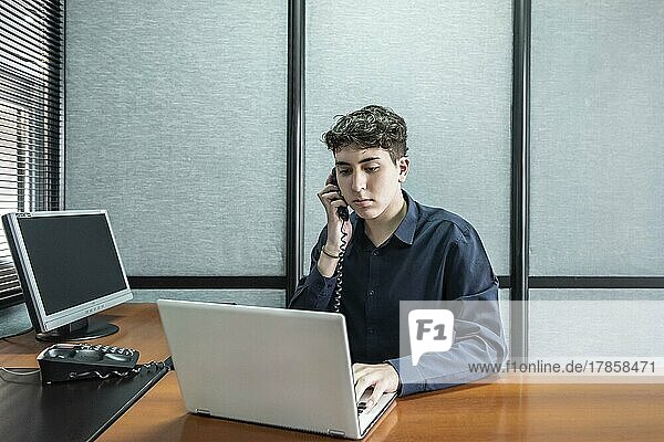 Junger Mann arbeitet an seinem Laptop  während er im Büro telefoniert