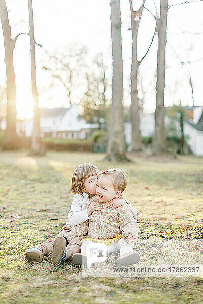 Toddler boy kisses baby sister on cheek while sitting in a field