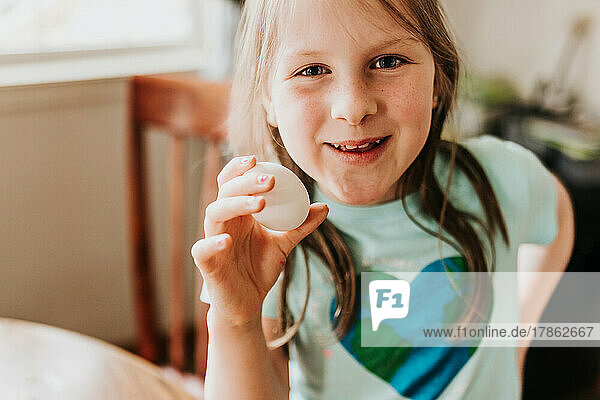Young smiling girl holds up white egg while looking at camera