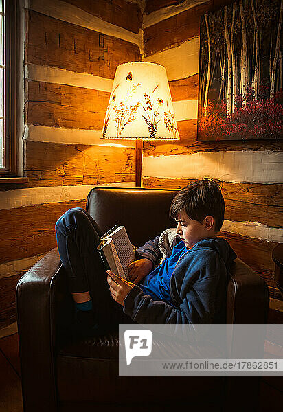 Boy reading book in chair in the corner of a rustic log cabin.