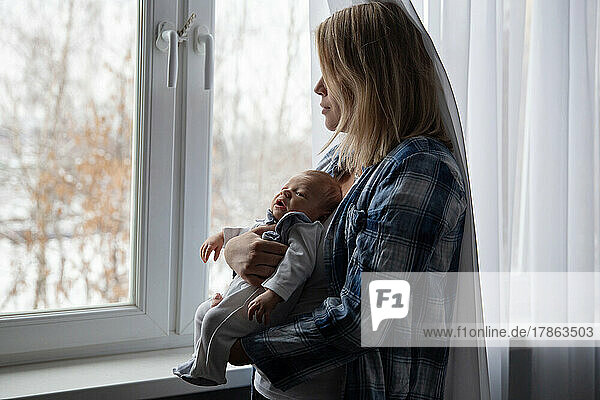 A young mother with her newborn baby in her arms stands by the window