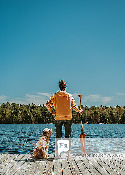Woman standing on dock with dog holding paddle looking out at lake.