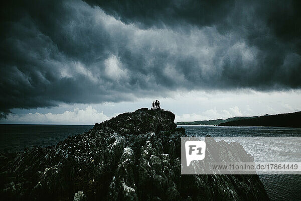 Group of people hike on rocky cliff over ocean with stormy sky