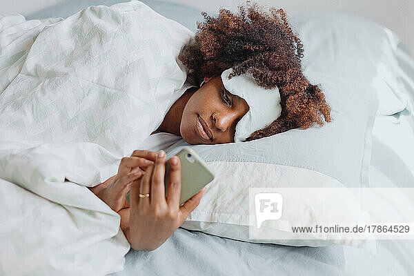 african woman with afrohair in bed with smartphone. sleepmask