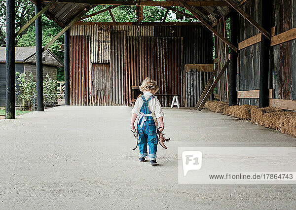 child in dungarees walking across a barn holding his toy dinosaurs