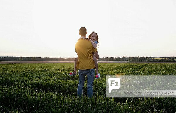 father carrying his daughter whilst walking in a field at sunset