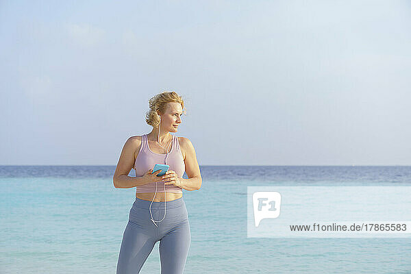 A sporty woman listens to music on her smartphone on the beach.