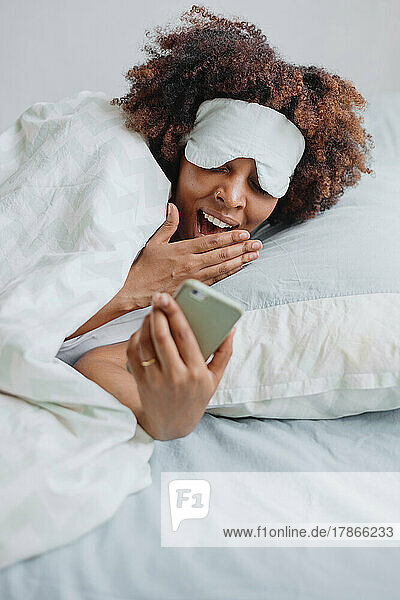 African woman yawns in bed and looks at messages on the phone