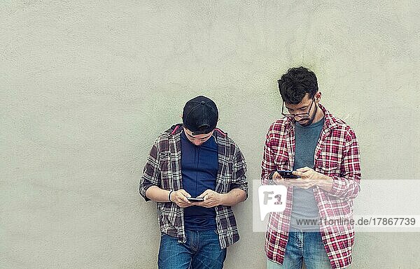 Two teenage friends on a wall checking their cell phones  Two friends leaning on a wall texting on their phones. Friend showing cell phone to his friend  Smiling friends checking cell phones