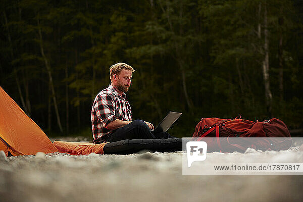 Businessman using laptop sitting on inflatable mattress at campsite