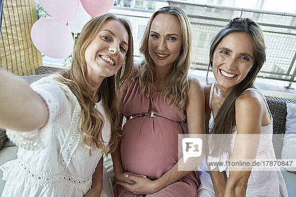 Happy friends taking selfie with pregnant woman sitting on sofa
