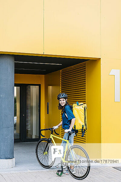 Smiling young delivery woman with bicycle standing in front of building
