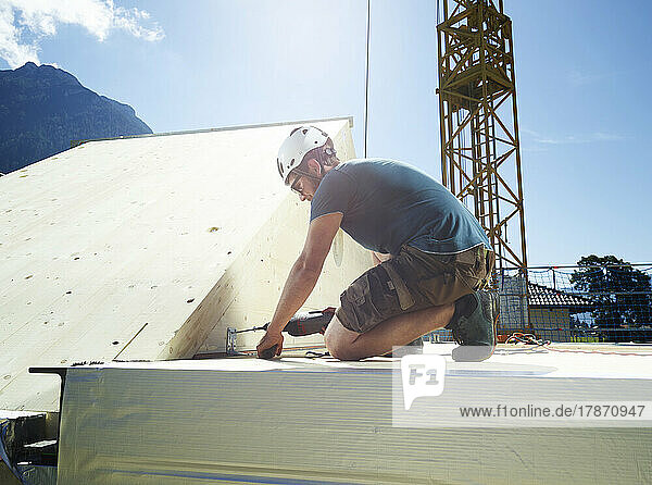 Junger Zimmermann installiert an einem sonnigen Tag auf der Baustelle ein Dach mit einer Bohrmaschine