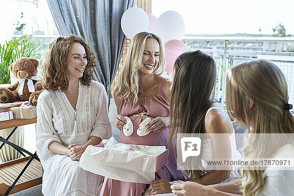 Happy pregnant woman holding baby booties sitting by friends on sofa