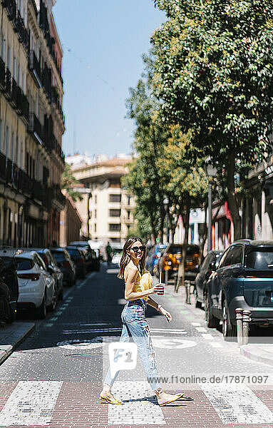 Young stylish woman with drink in hand crossing street
