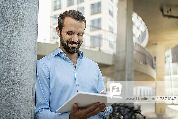 Lächelnder junger Geschäftsmann mit Tablet-PC  der unter einer Brücke steht