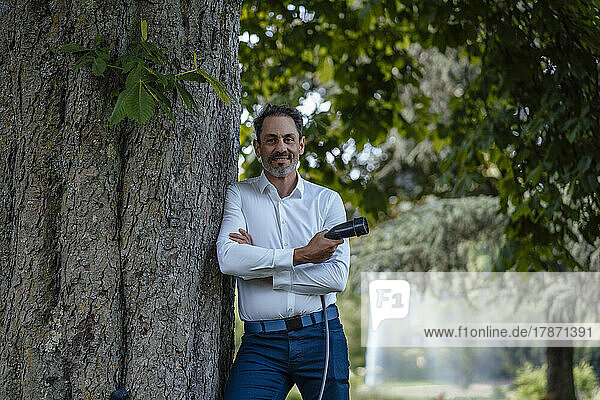 Mature businessman with arms crossed holding charging cable leaning on tree trunk