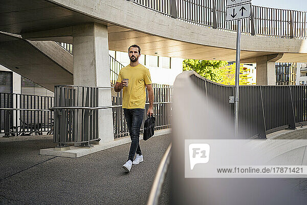 Young businessman with coffee cup and bag walking on footbridge