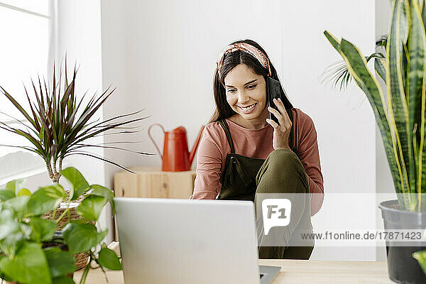 Smiling woman with laptop talking on phone sitting by houseplants at home