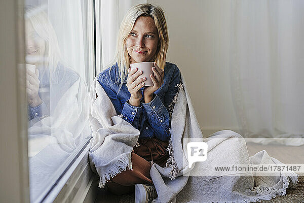Young woman wrapped in blanket holding coffee cup sitting by window at home