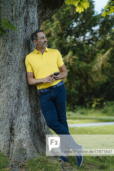 Smiling mature man holding smart phone leaning on tree trunk
