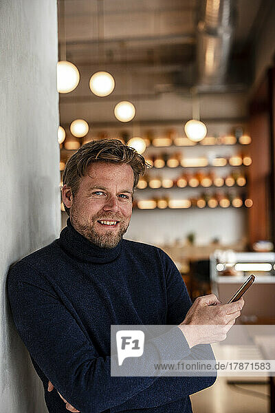 Smiling businessman with mobile phone leaning on wall in cafe