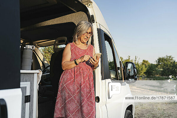 Mature woman using mobile phone leaning at doorway of motor home