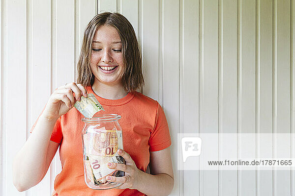 Smiling girl holding glass jar with European currency in front of wall