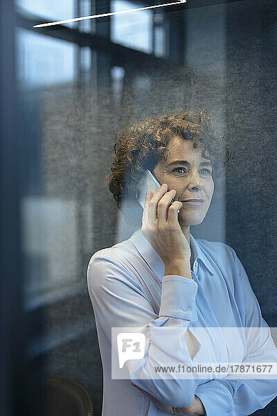Smiling businesswoman talking on smart phone seen through glass