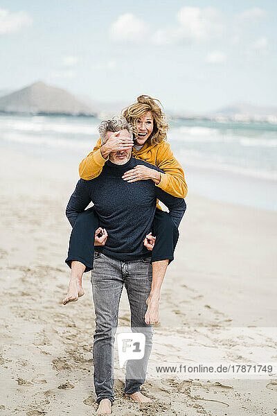 Fröhliche Frau bedeckt die Augen eines Mannes mit der Hand und genießt die Huckepackfahrt am Strand