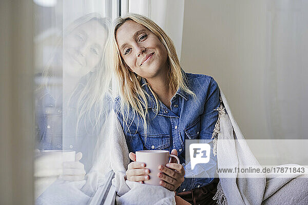 Smiling woman with coffee cup leaning on glass window at home