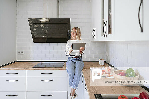 Thoughtful woman with tablet PC sitting on kitchen counter at home