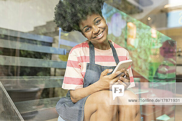 Glückliche junge Frau mit Smartphone sitzt im Café und sieht durch die Glasscheibe