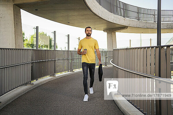 Businessman with disposable coffee cup and bag walking on footbridge