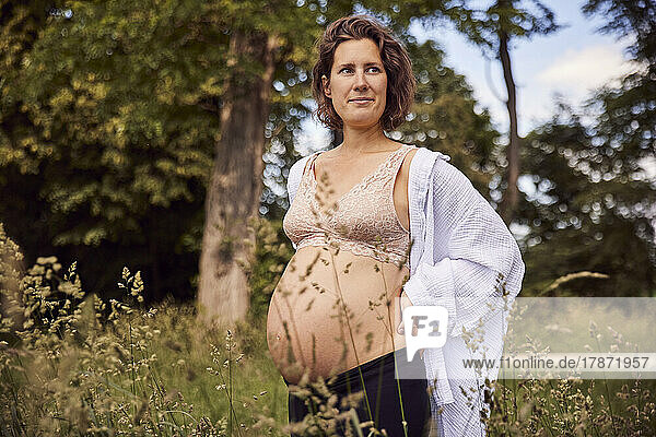 Smiling pregnant woman standing with hand on hip in grass