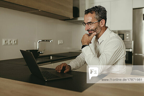 Lächelnder Geschäftsmann mit der Hand am Kinn und Laptop zu Hause