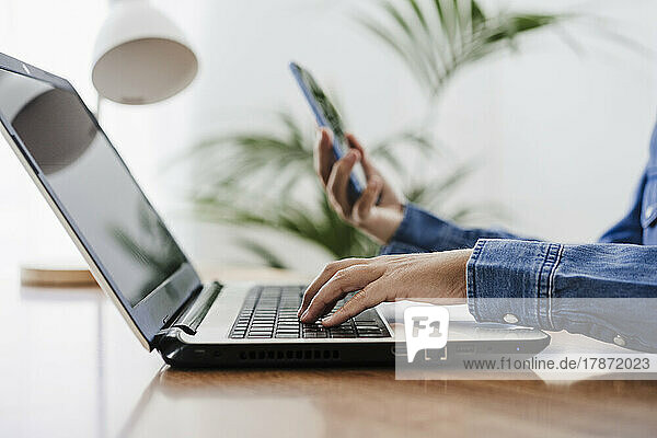 Freelancer using laptop holding smart phone at home office