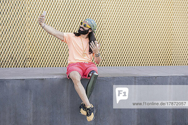 Glücklicher Mann mit Beinprothese  der ein Selfie mit dem an der Wand sitzenden Smartphone macht