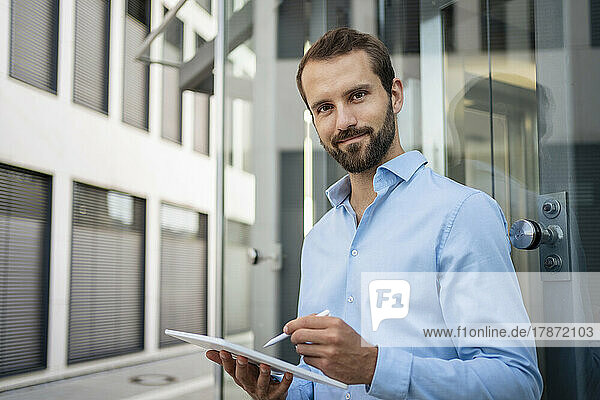 Smiling businessman with tablet PC standing in front of glass
