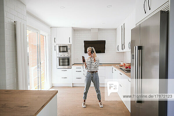 Woman with mobile phone listening music on headphones in kitchen at home