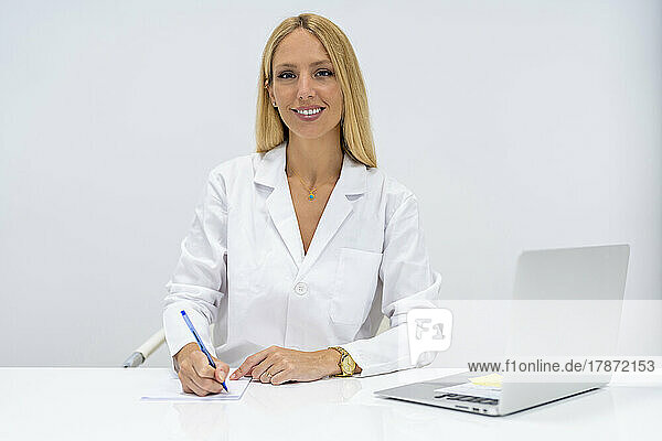 Smiling healthcare worker with note pad and laptop sitting at desk in clinic
