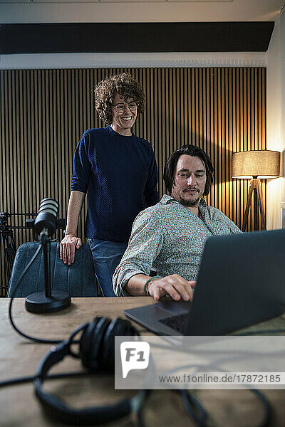 Smiling presenter talking with colleague using laptop in recording studio