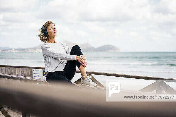 Mature woman wearing wireless headphones sitting on railing at beach