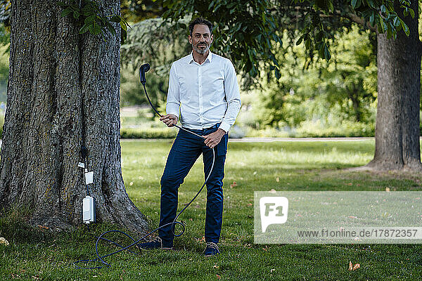 Mature businessman holding charging cable standing by tree