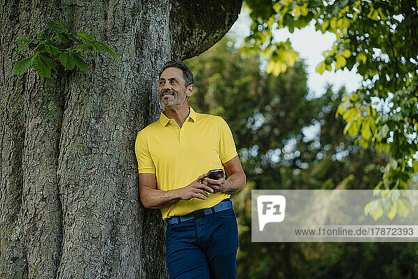 Smiling man holding smart phone leaning on tree trunk