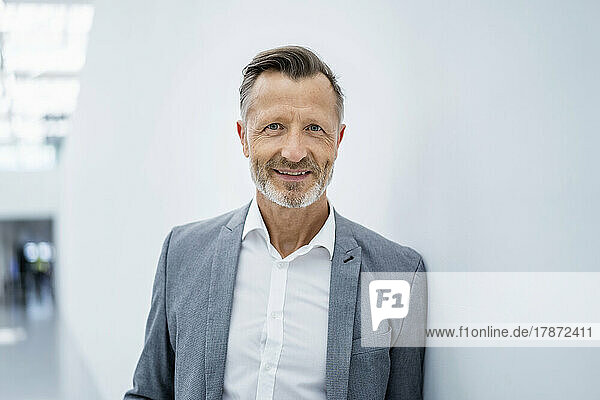Smiling mature businessman in blazer leaning on white wall