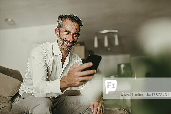 Lächelnder reifer Geschäftsmann  der zu Hause auf dem Sofa sein Mobiltelefon benutzt